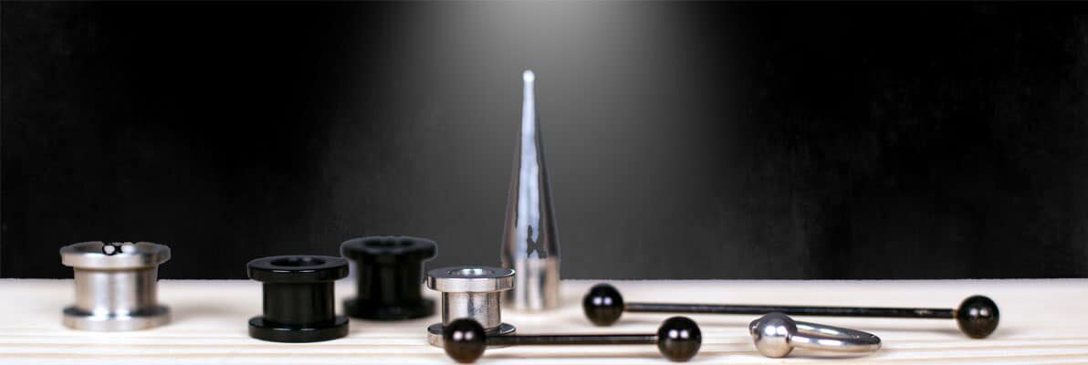 Assorted body piercing jewelry, including barbells, plugs, and a taper, displayed against a black background.