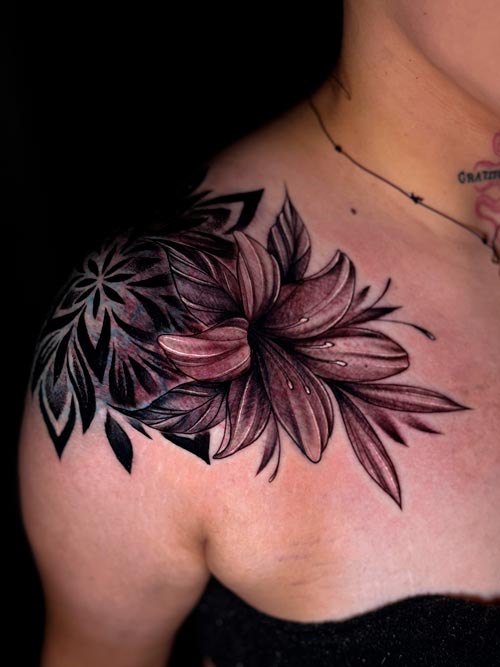 Close-up of a custom floral tattoo on the upper chest and shoulder, featuring a large lily with fine shading, soft black and grey tones, and bold geometric blackwork accents.